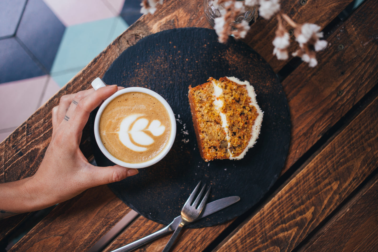 Hand holding cup of latte made with freshly roasted Peruvian coffee next to slice of carrot cake on black plate