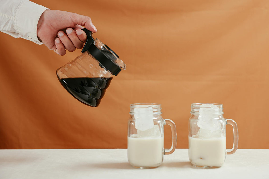 Hand pouring coffee into ice-filled milk glasses against a warm background.