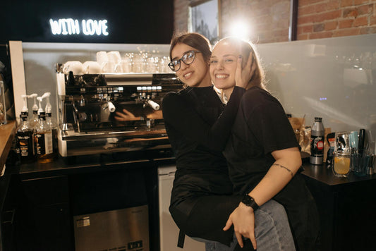 Two smiling baristas in black attire embrace warmly inside a cozy coffee shop with a modern espresso machine.