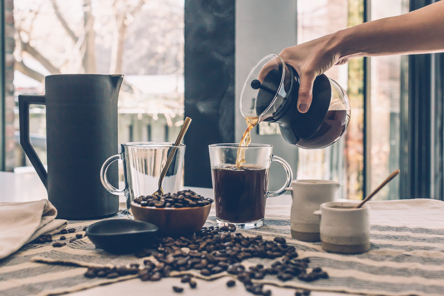 Pouring brewed specialty Kenyan coffee online into glass mugs with coffee beans and ceramic containers on table