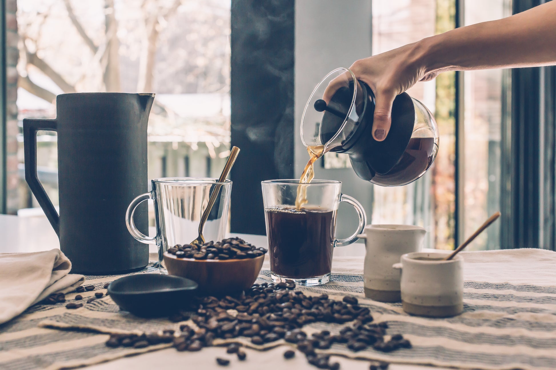 Pouring brewed specialty Kenyan coffee online into glass mugs with coffee beans and ceramic containers on table
