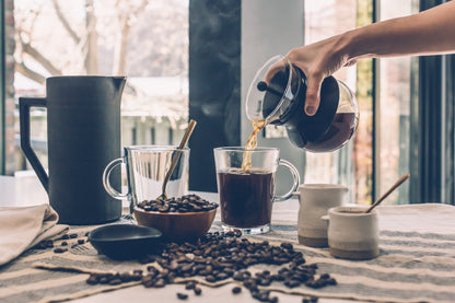 Pouring brewed specialty Kenyan coffee online into glass mugs with coffee beans and ceramic containers on table